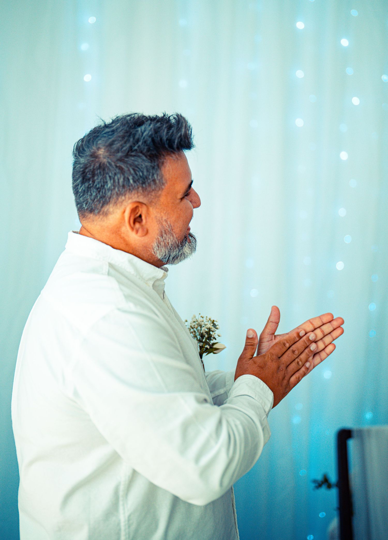 A person in a white suit with a boutonniere stands in profile against a light blue background with sparkles.