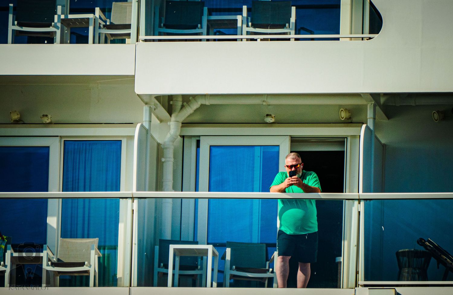 A person in a teal shirt stands on a cruise ship balcony against blue cabin windows.
