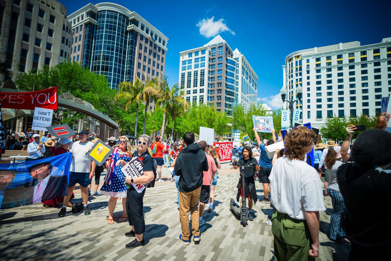 Protesters gather in front of modern office buildings during a downtown demonstration with signs and banners.