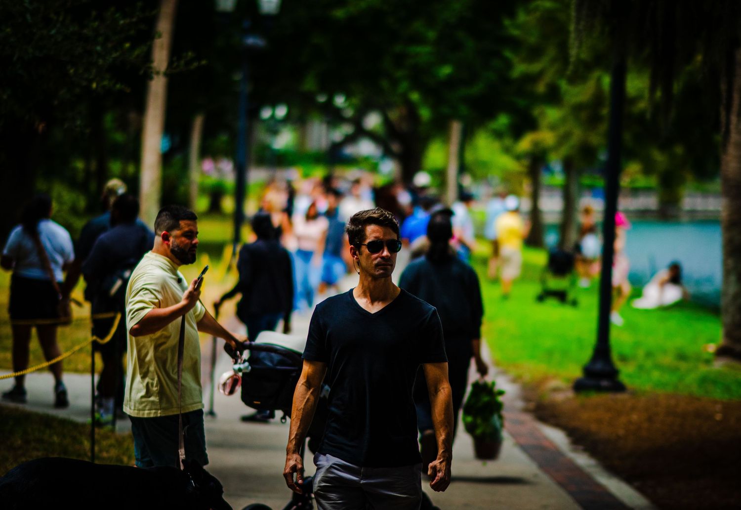 Man in black t-shirt and sunglasses walks along a busy park pathway surrounded by a crowd of people on a sunny day.