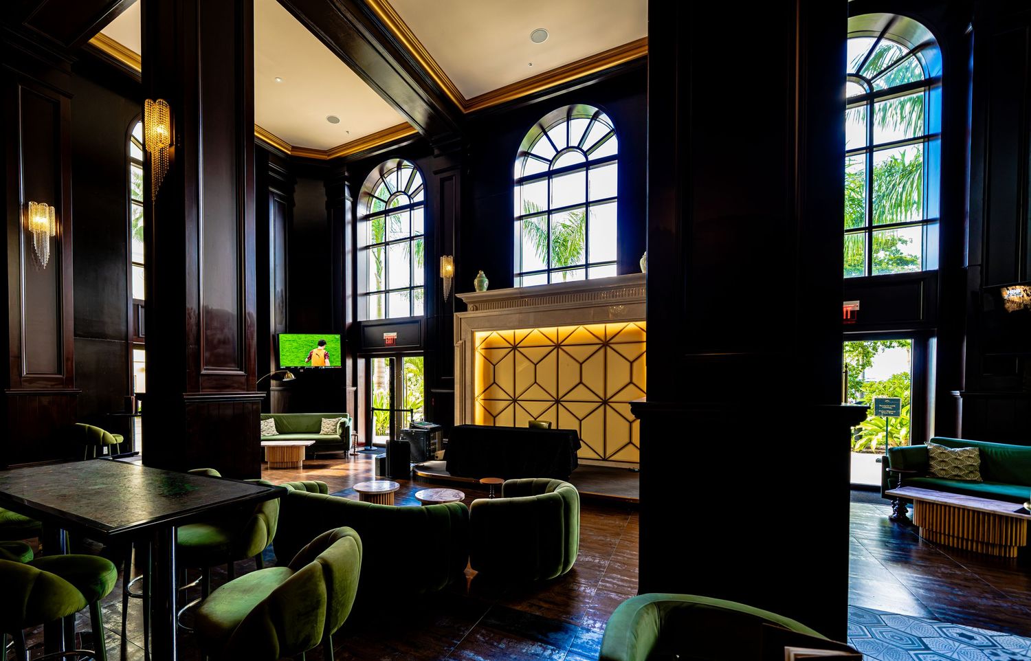 Elegant hotel lobby with dark wood paneling, arched windows, green velvet chairs, and a gold-lit reception desk.