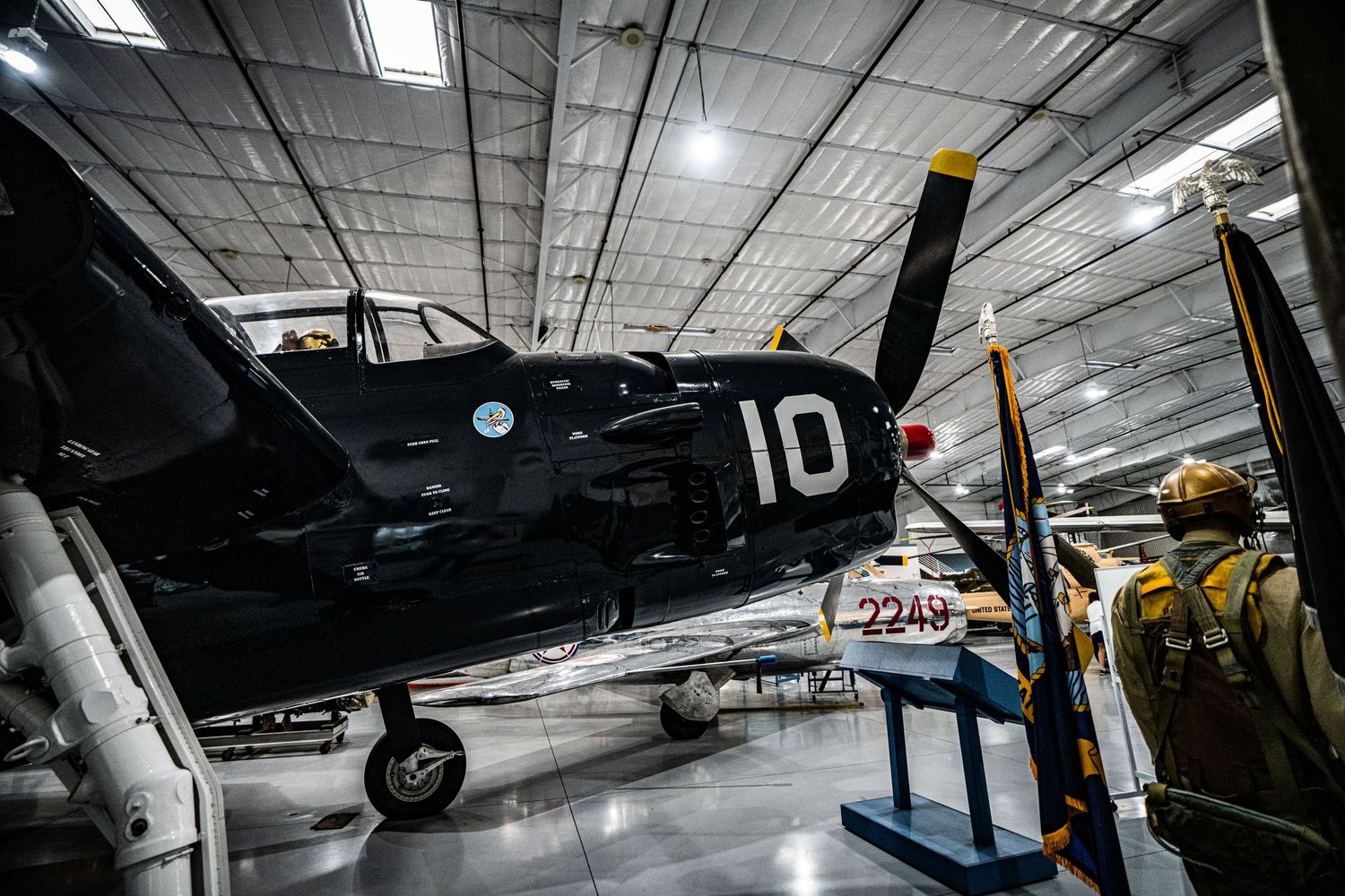 Vintage WWII Navy fighter plane numbered 10 displayed in aviation museum hangar with visitors nearby.