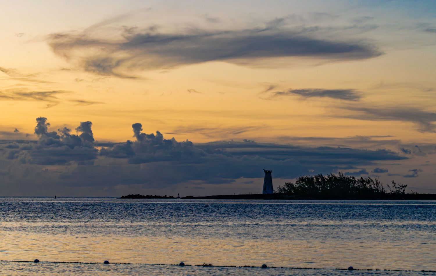 Silhouette of a lighthouse at sunset over calm ocean waters with dramatic clouds in warm orange and blue hues.