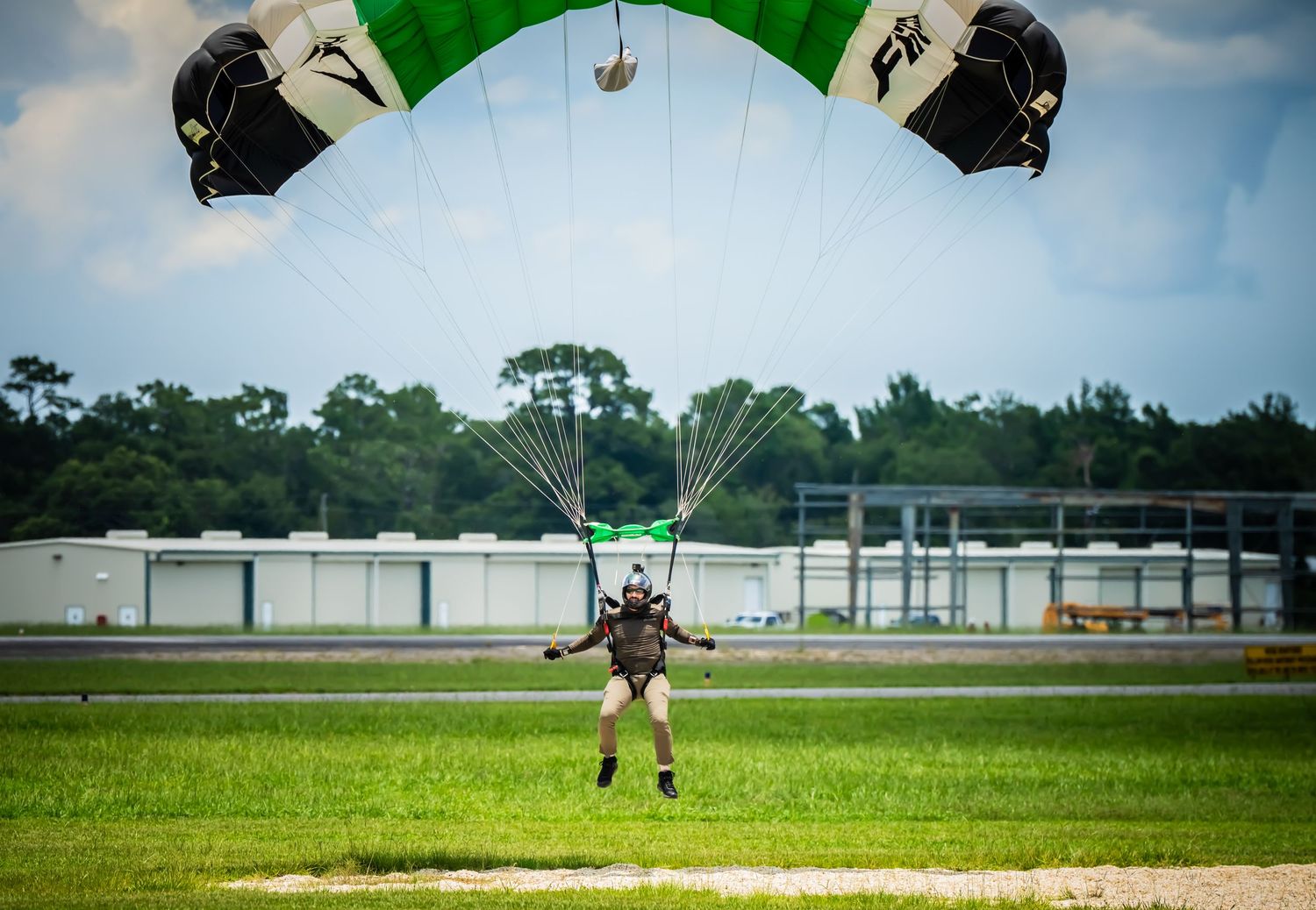 Skydiver with green parachute landing on grassy field near storage units.