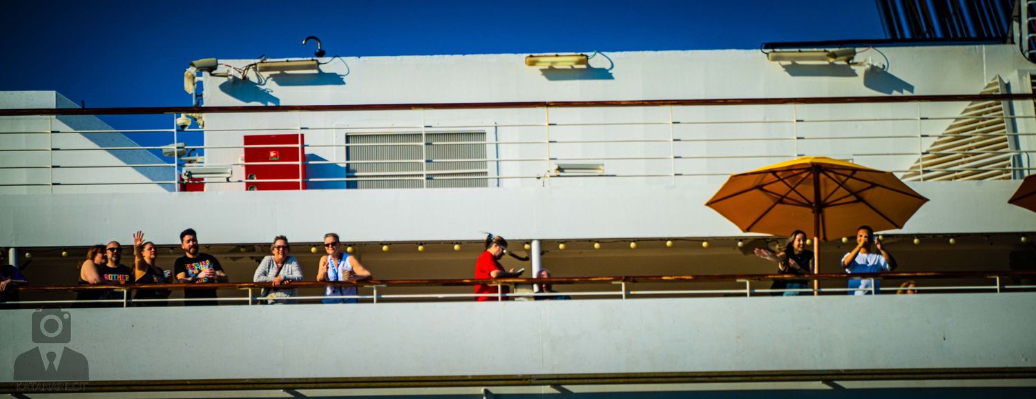 Spectators line the upper deck railing of a cruise ship watching an event or activity below.