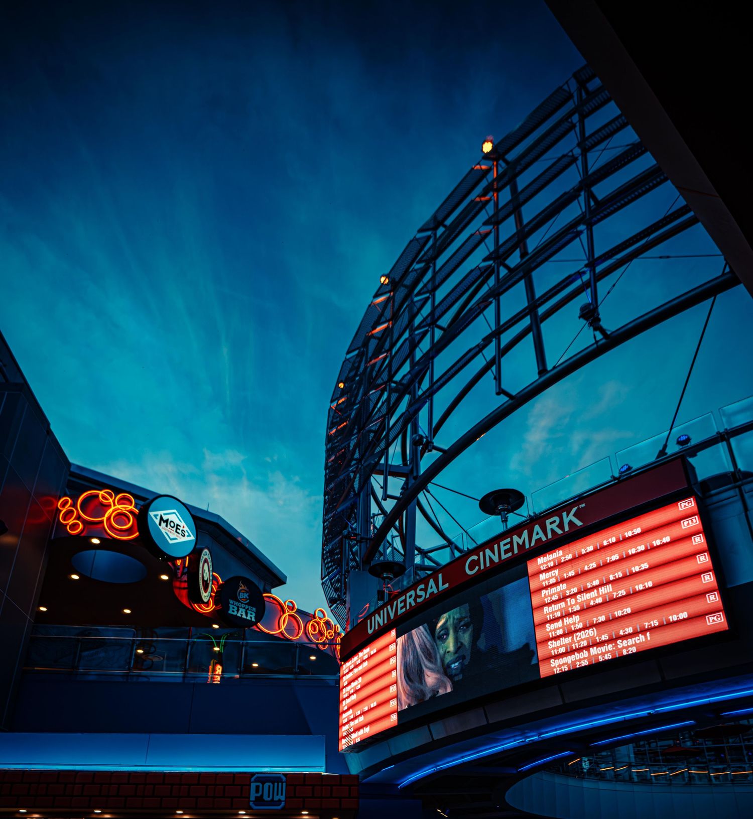Neon signs and electronic displays illuminate a dark urban street with a dramatic blue twilight sky.