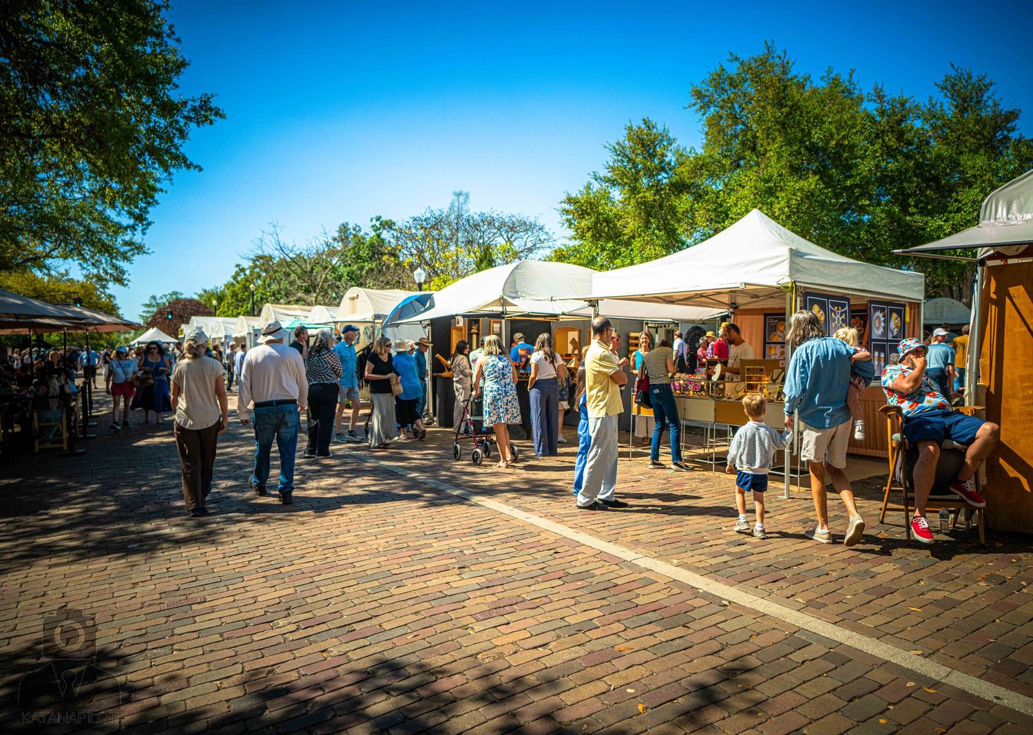 Outdoor farmers market with white vendor tents and shoppers browsing local goods on a sunny summer day.