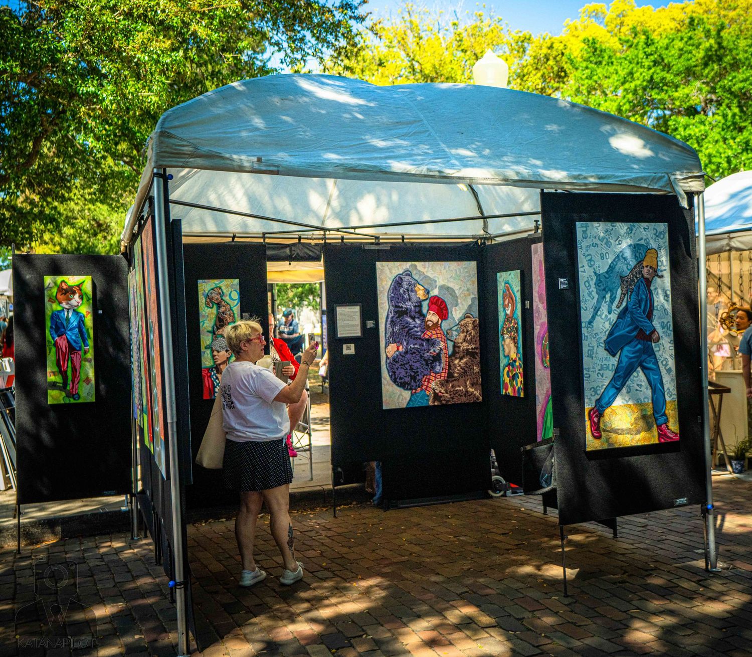 Art vendor booth displays colorful stained glass artwork at an outdoor festival under a blue canopy tent.
