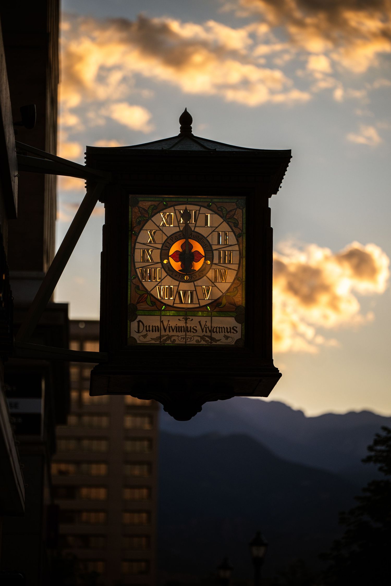 Ornate illuminated street clock with Latin inscription at sunset, mountains and dramatic clouds in background.