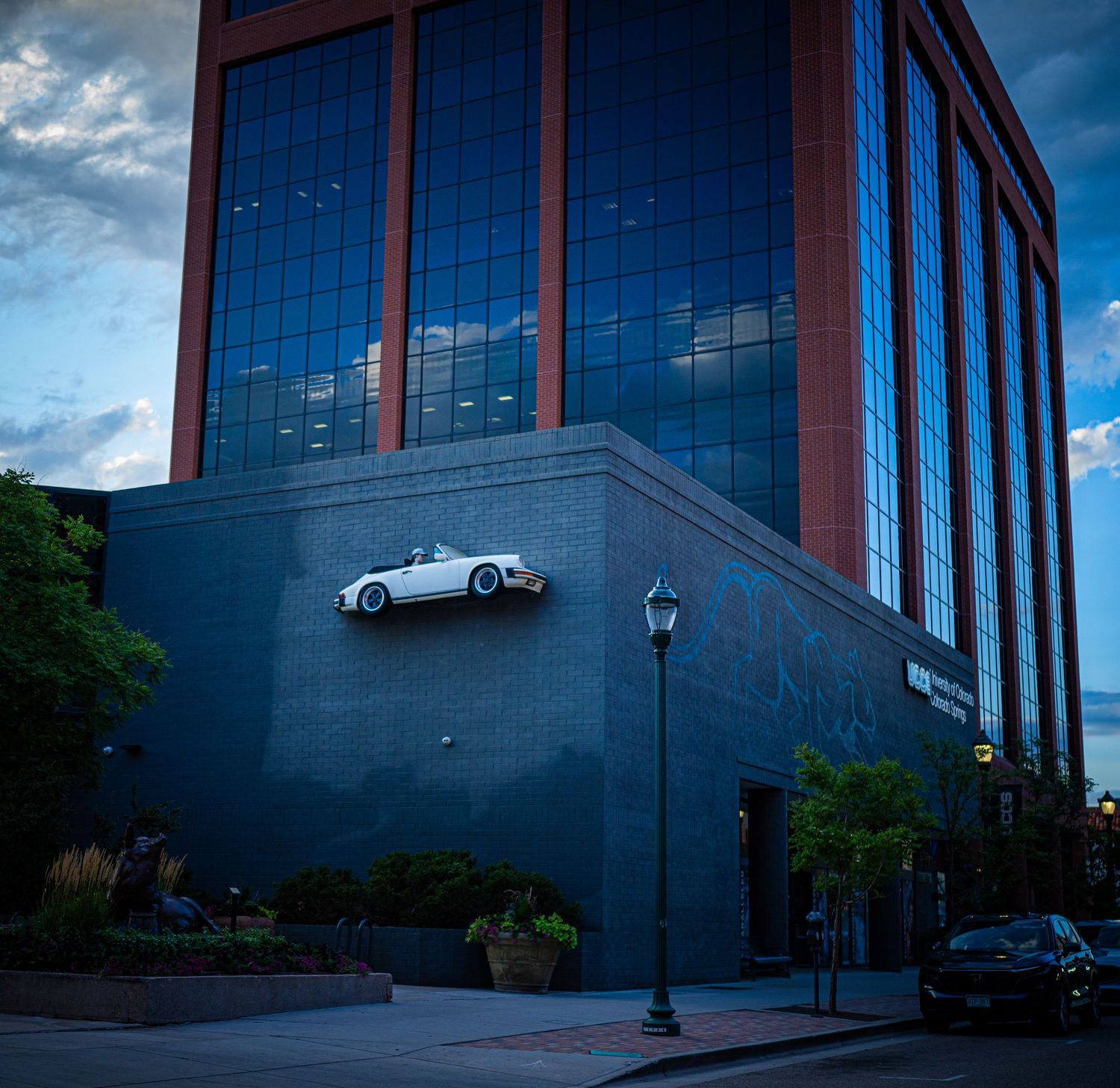 A white sports car mounted on the exterior wall of a modern brick and glass building at dusk.