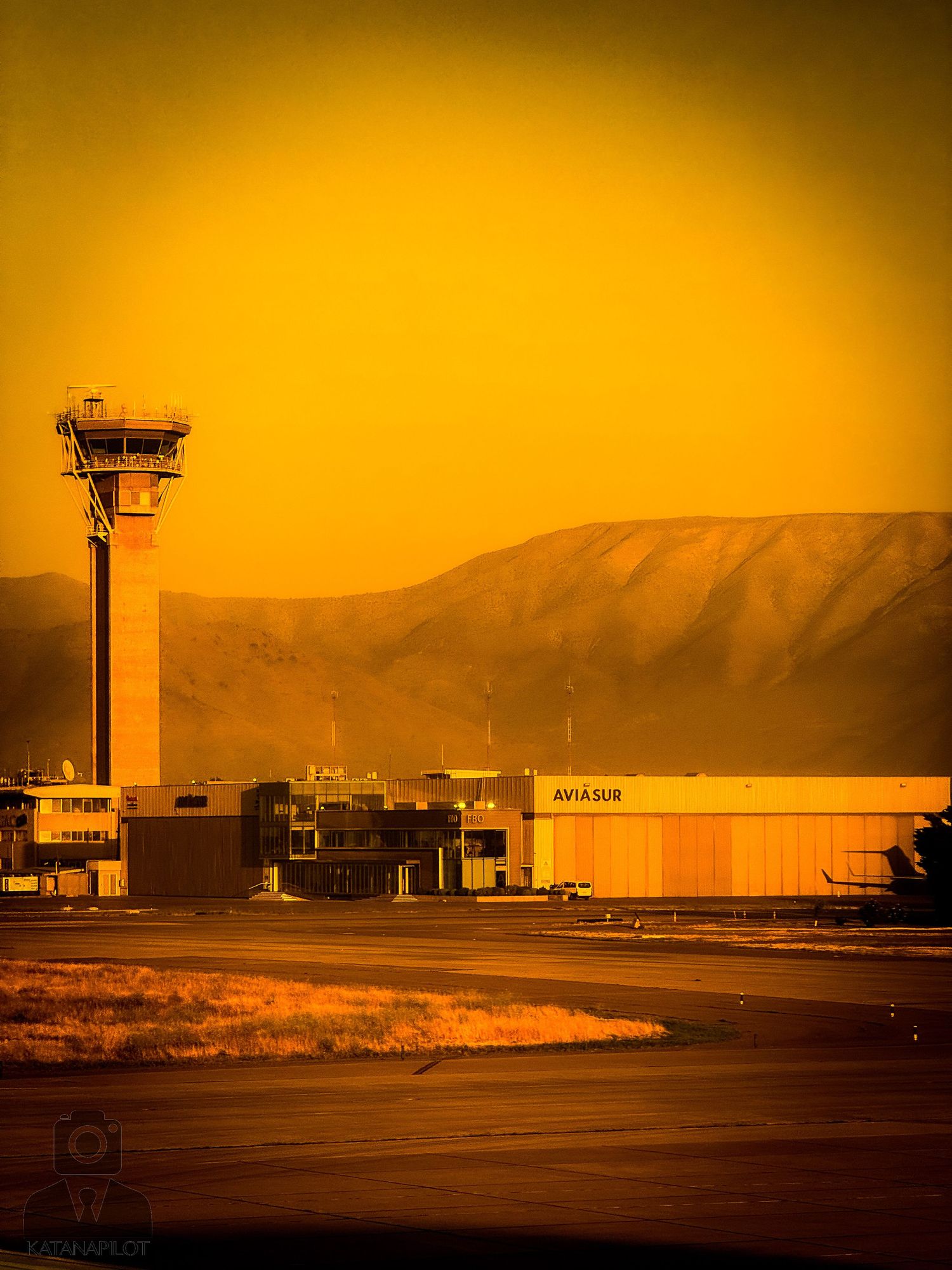 Airport control tower and runway at sunset with orange sky and mountain silhouettes.