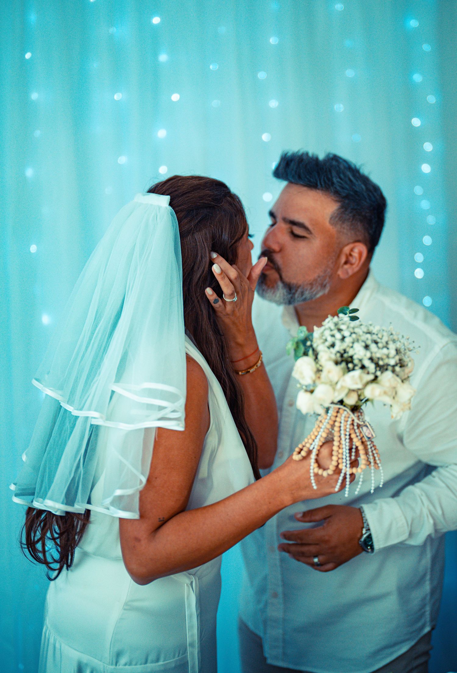 A romantic wedding moment captured against a turquoise backdrop with twinkling lights and white floral bouquet.
