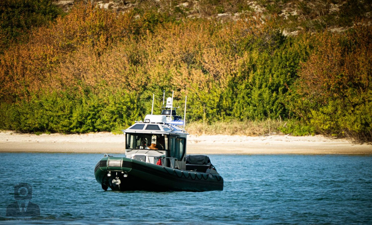 A police or patrol boat cruises along coastal waters near a beach with green foliage in the background.