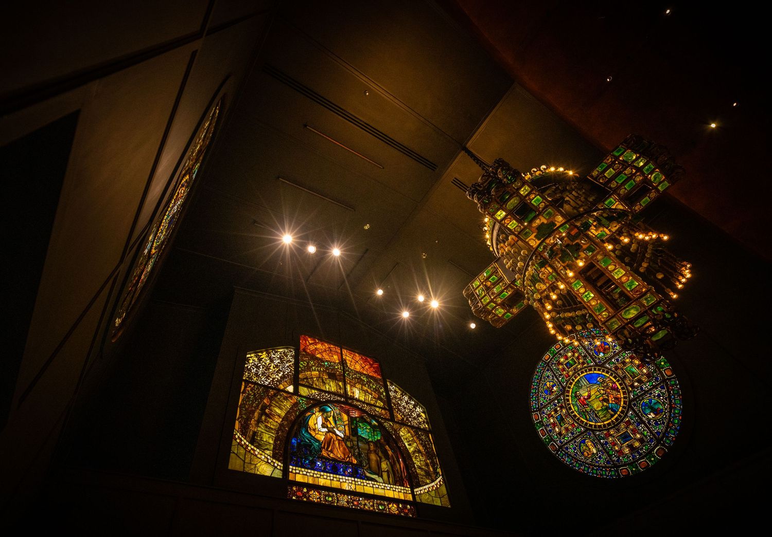 Ornate stained glass ceiling fixtures and chandeliers illuminated in a dark cathedral-like space.