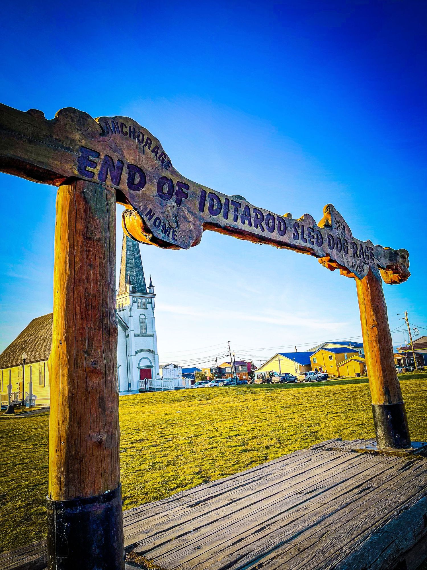 Wooden Iditarod trail marker sign with church steeple visible in the background.