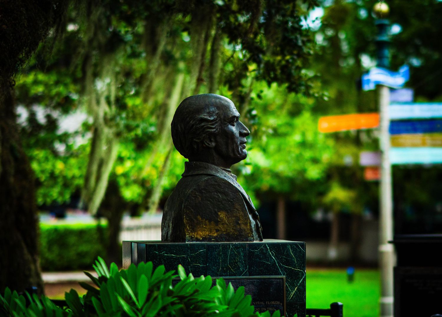 Bronze bust sculpture in a lush green park with colorful signs in the background.