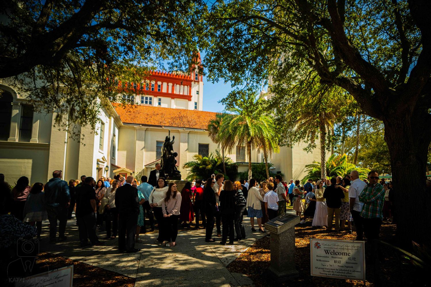 Crowd gathered under shady trees in courtyard of Spanish colonial style building.