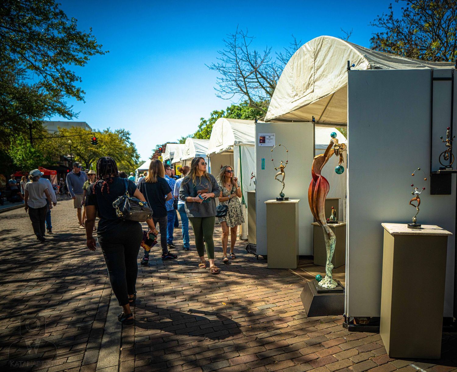 Row of white exhibition tents with artwork displays and visitors walking along a shaded path.