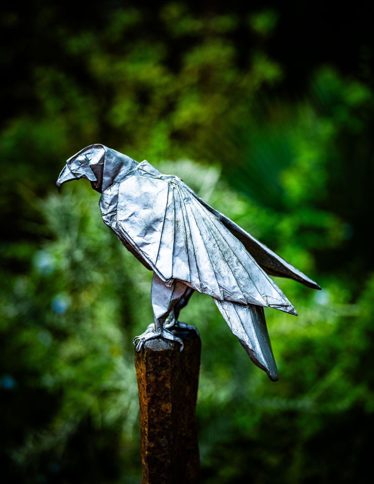 Metallic origami bird sculpture perched on a wooden post with blurred natural background.