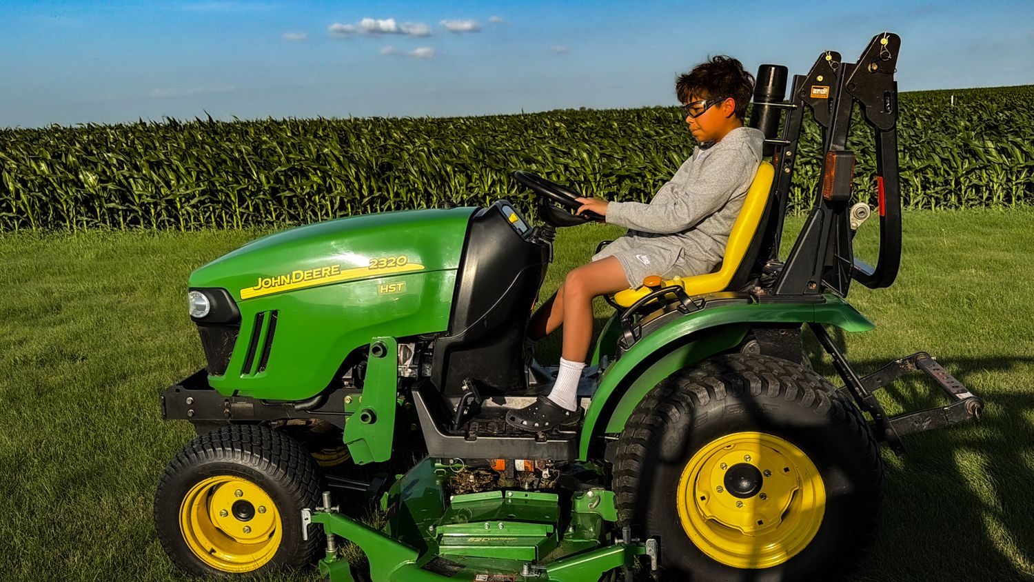 Person riding a green John Deere compact tractor on a farm with tall corn crops in the background.
