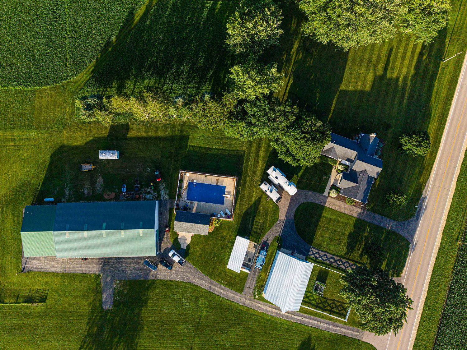 Aerial drone view of a rural farm property with green fields, blue metal barn, outbuildings, and tree-lined gravel driveway.