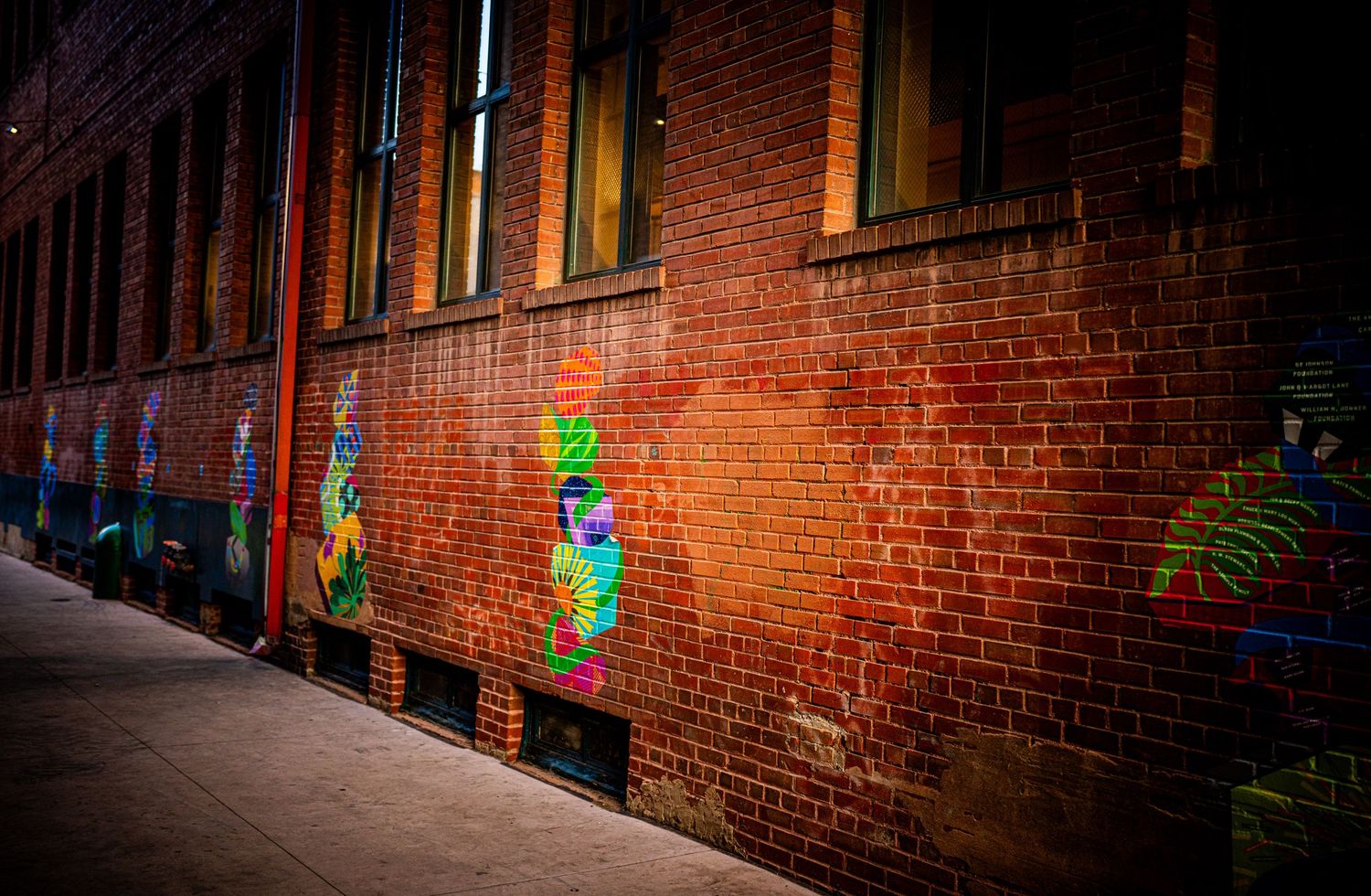 Colorful graffiti murals on a red brick building wall in an urban alley with a sidewalk.