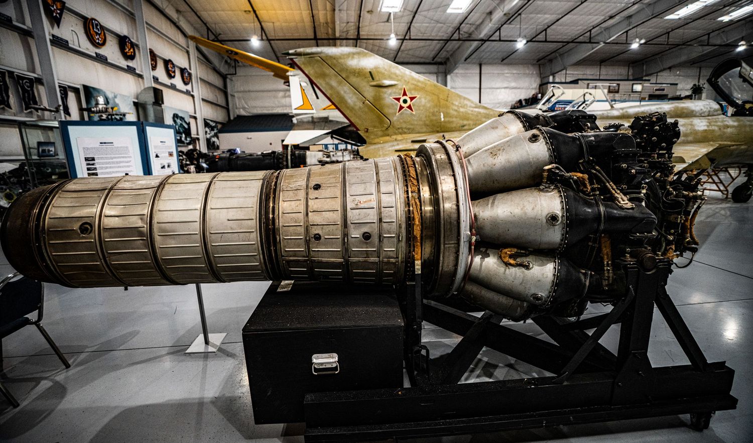 Large jet engine on display in an aviation museum hangar with aircraft and memorabilia in the background.