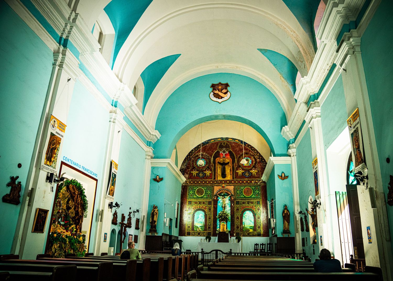 Interior of a Catholic church with turquoise vaulted ceiling, white arches, and ornate golden altar at the front.