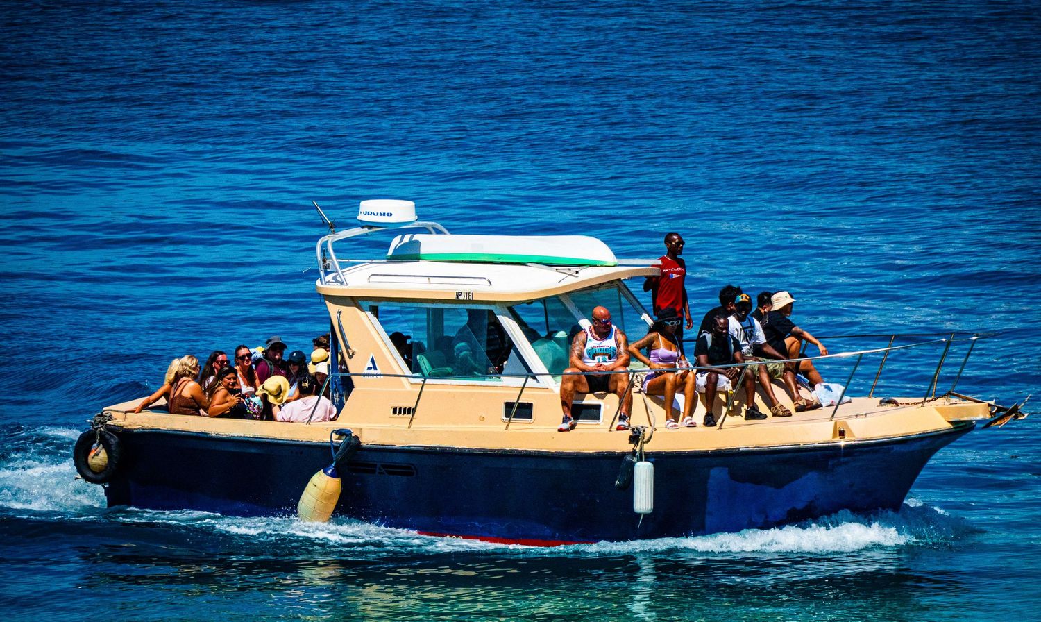A group of tourists enjoying a boat tour on a blue motorboat cruising through clear turquoise Mediterranean waters.