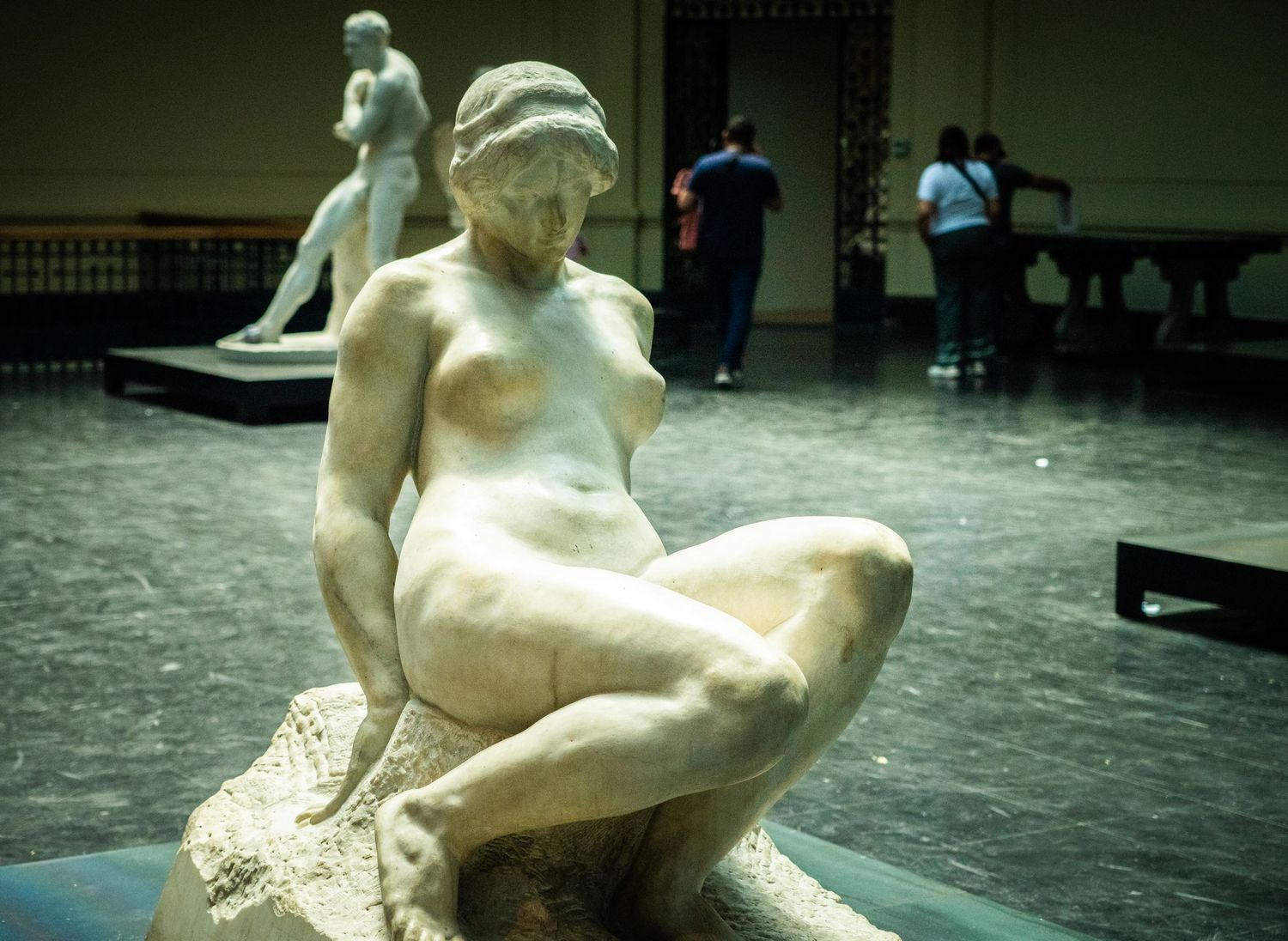 Classical marble sculpture of a nude figure seated on a rock in an art gallery setting.