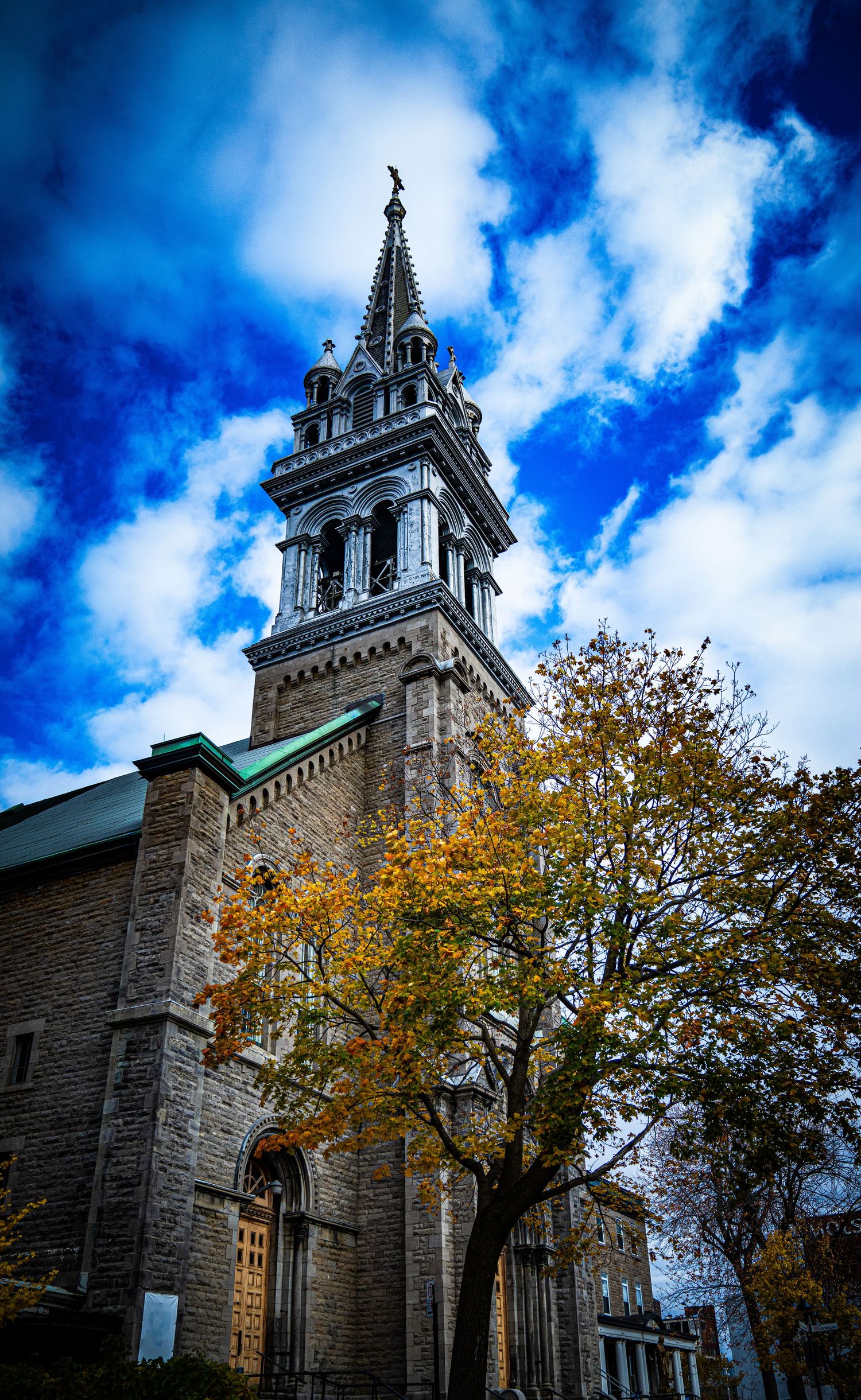 Historic stone church with spire and autumn trees against dramatic blue sky with white clouds.