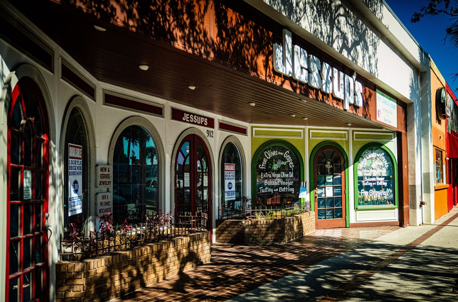 A panoramic view of a shopping mall's exterior with arched walkways and storefronts casting shadows on the ground.