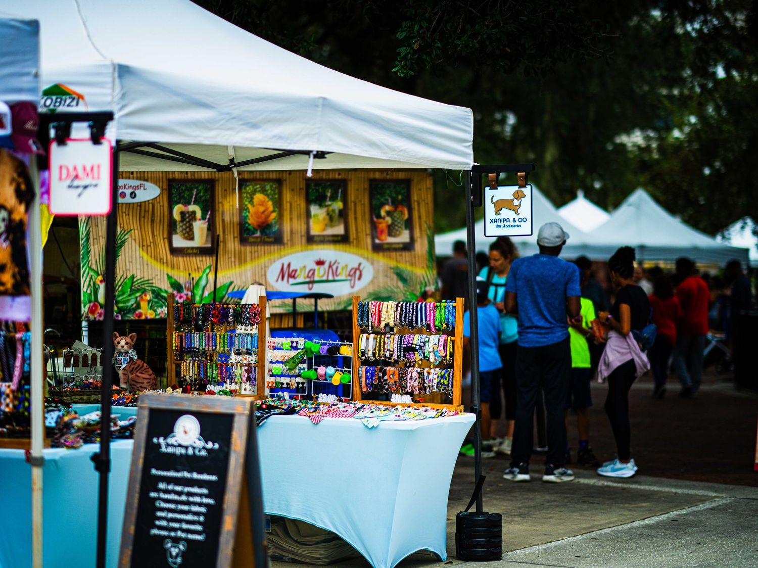Colorful outdoor market vendor booth with crafts under white tent at busy street fair festival.