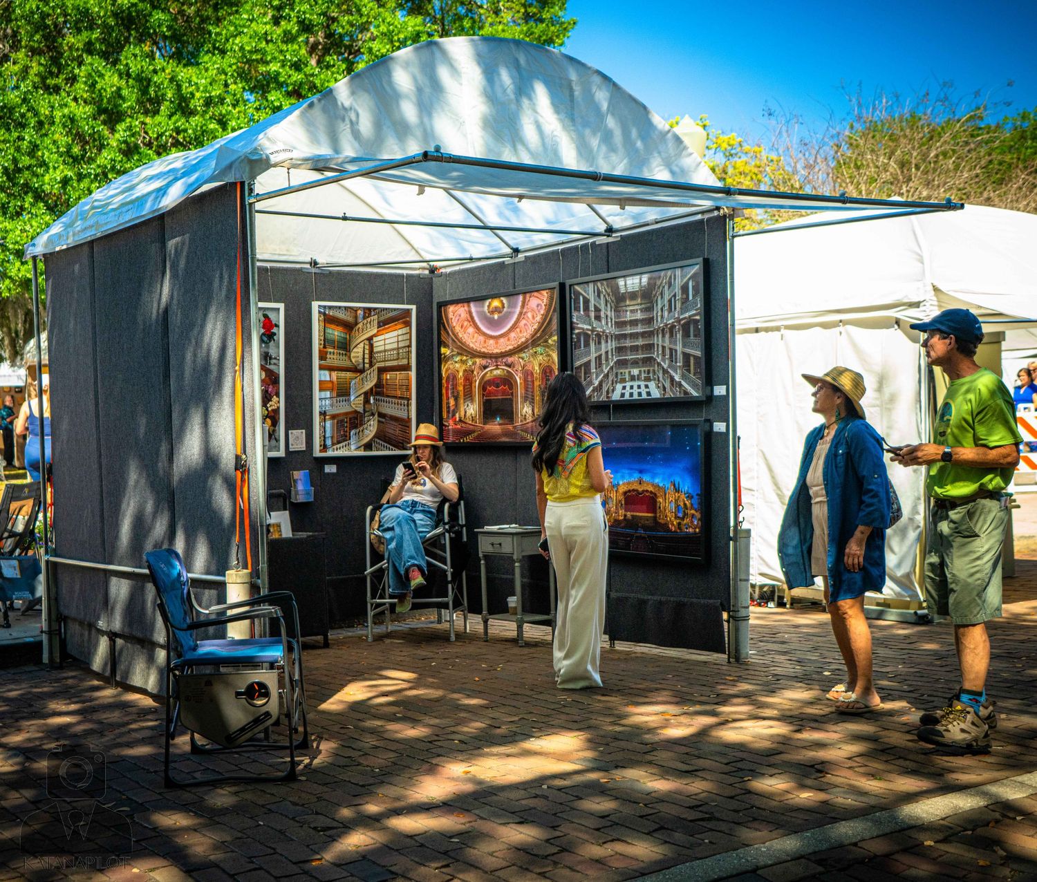 Outdoor art gallery setup with white canopy tents showcasing artwork displays under bright blue sky.