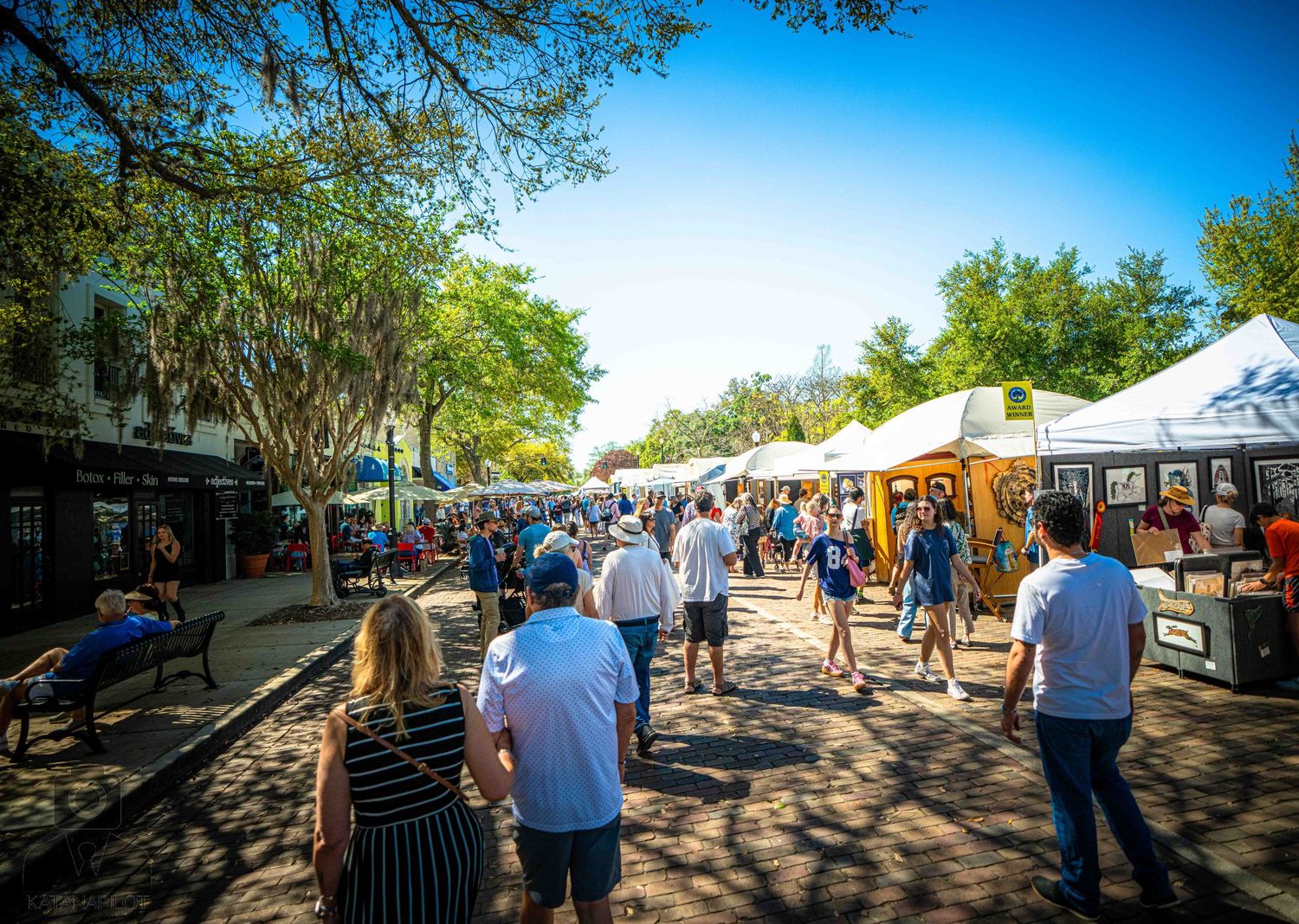 People browse vendor stalls and tents at an outdoor market on a sunny day with blue skies overhead.
