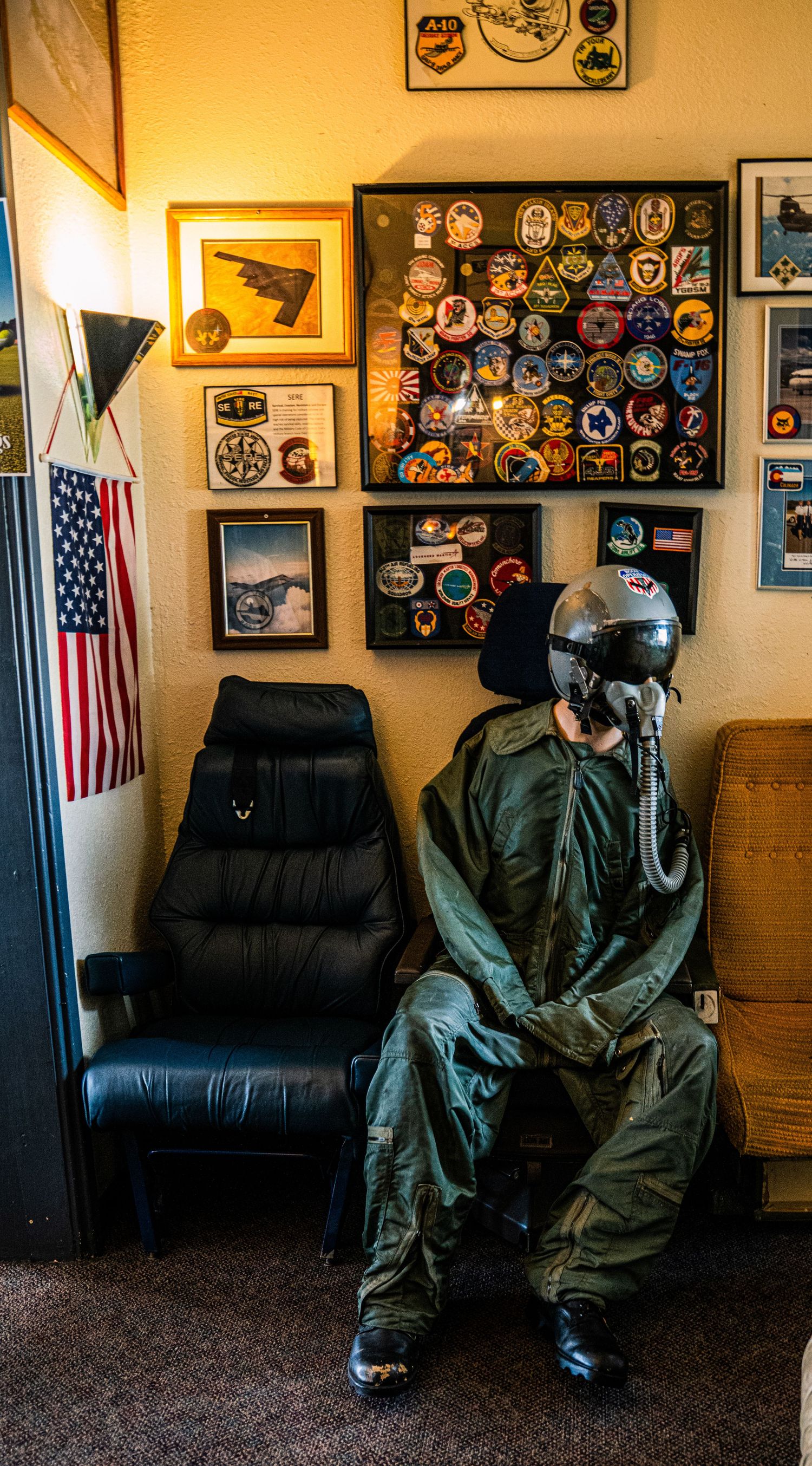 Military flight suits displayed on chairs in a room decorated with patches and American flags on the walls.
