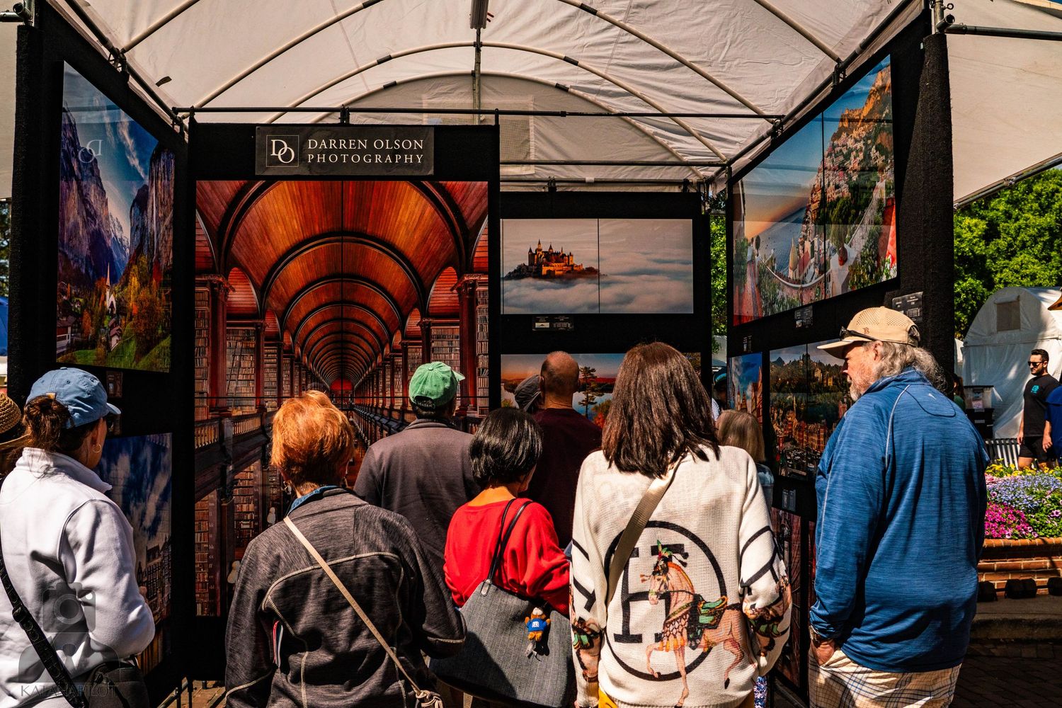 Art gallery booth with photography displays drawing crowd of visitors under white canopy.