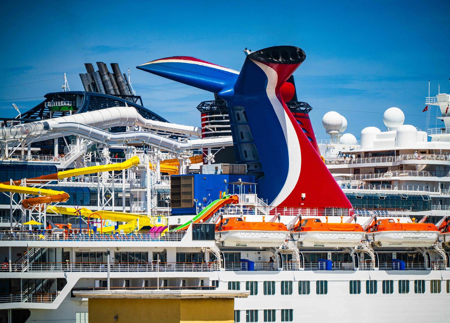Carnival cruise ship with colorful funnel, water slides, and lifeboats docked at port under blue sky.