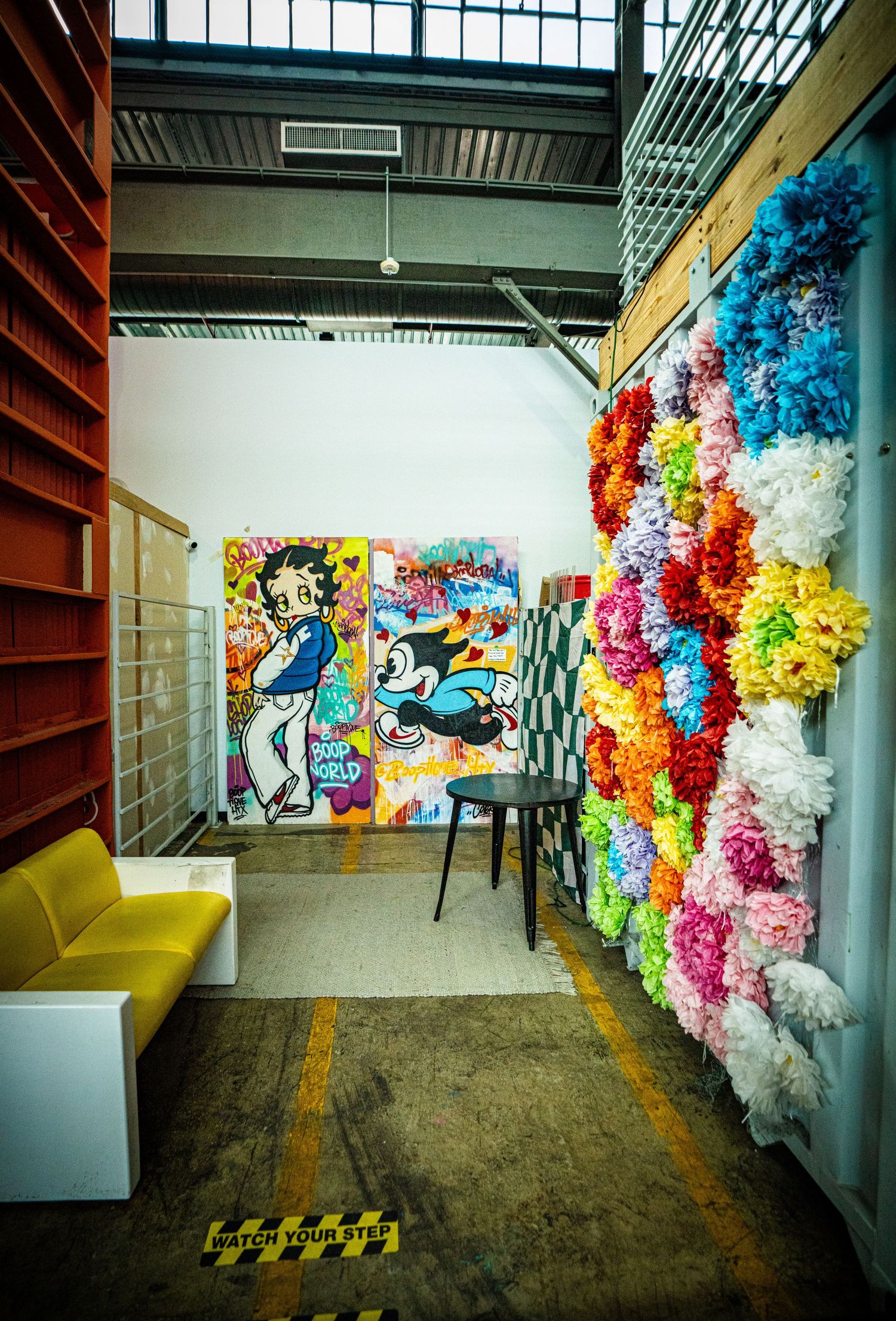 Eclectic indoor art space featuring Betty Boop graffiti mural, rainbow floral wall installation, and yellow seating in a warehouse.