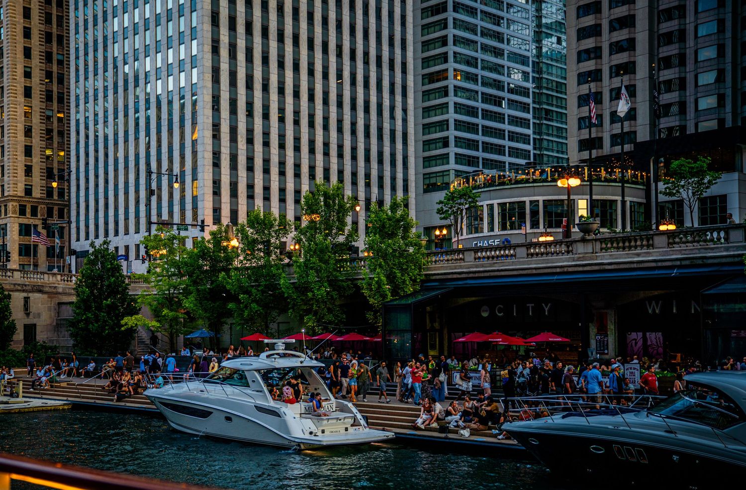 Chicago Riverwalk at sunset with boats cruising along the river between illuminated skyscrapers.