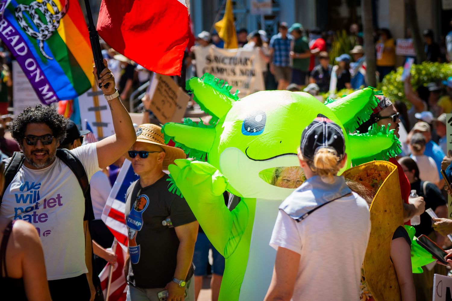 Parade participants march with colorful pride flags and inflatable green dinosaur during sunny outdoor celebration.