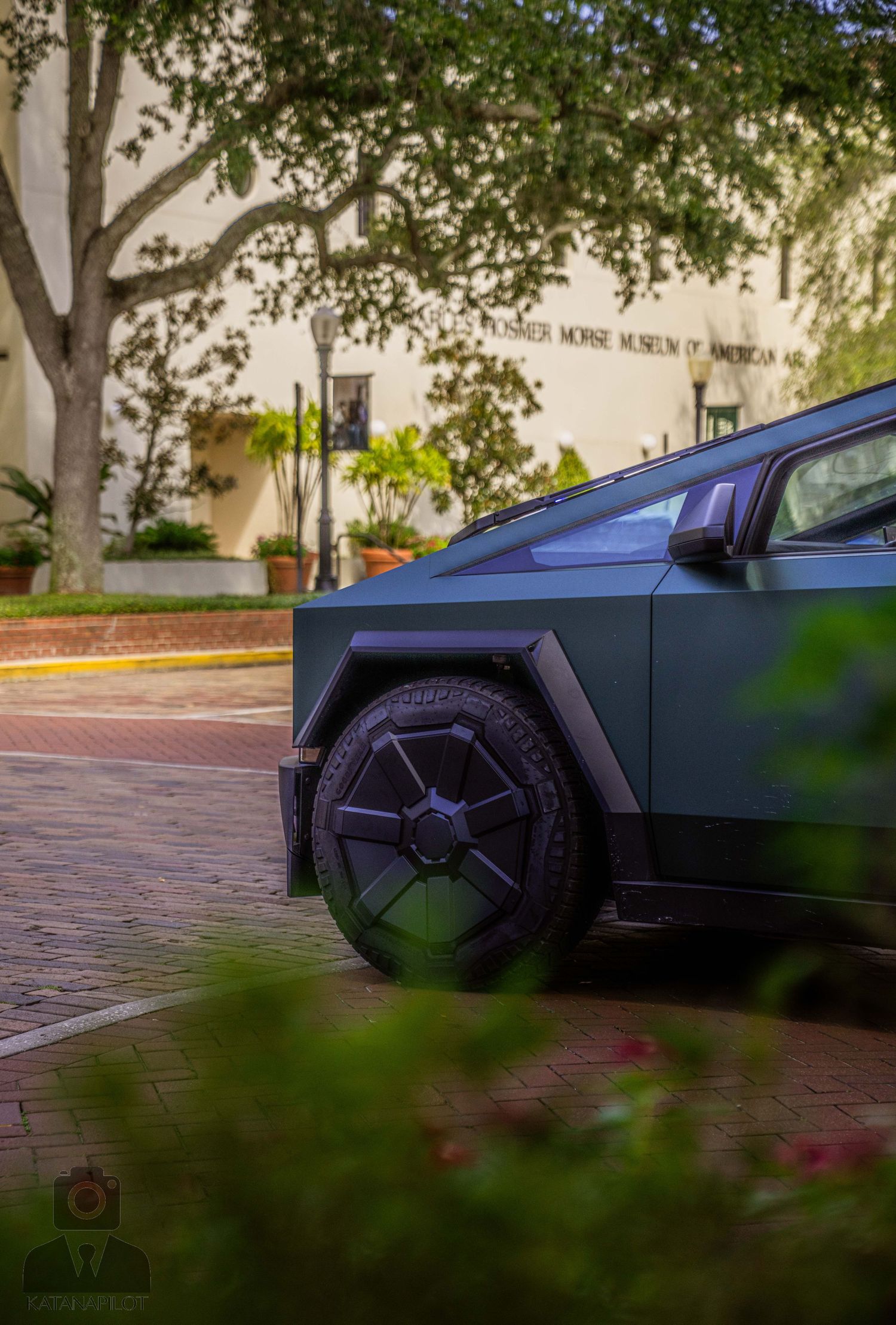 Low angle view of a dark car parked under trees on brick pavement.