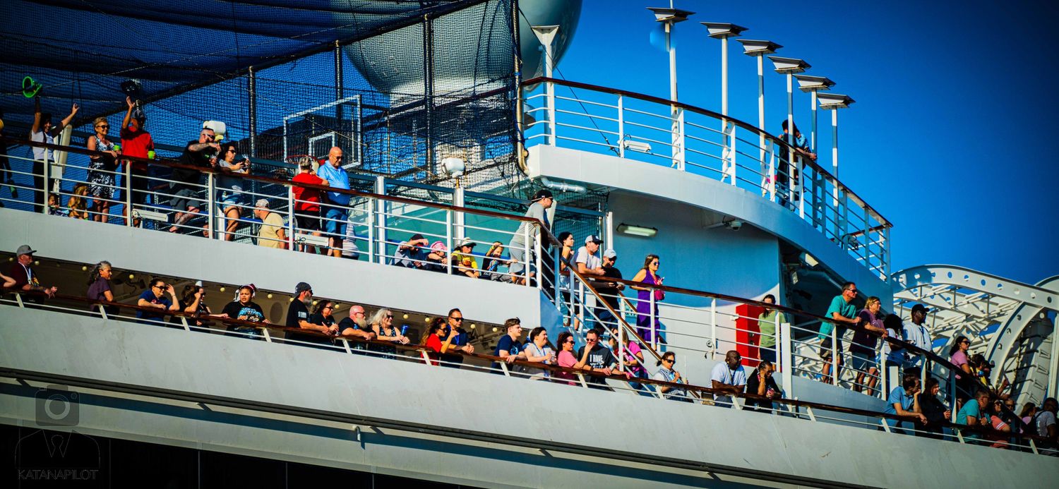 Passengers relax on the upper deck of a cruise ship against a bright blue sky on a sunny day.