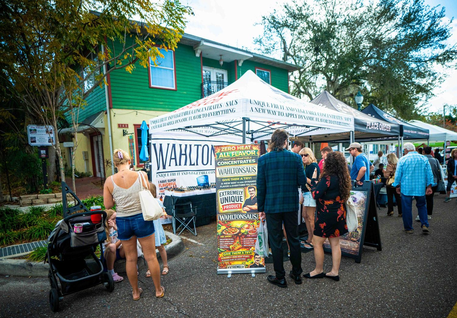 Outdoor farmers market stalls with vendors and shoppers browsing local produce and goods on a sunny day.