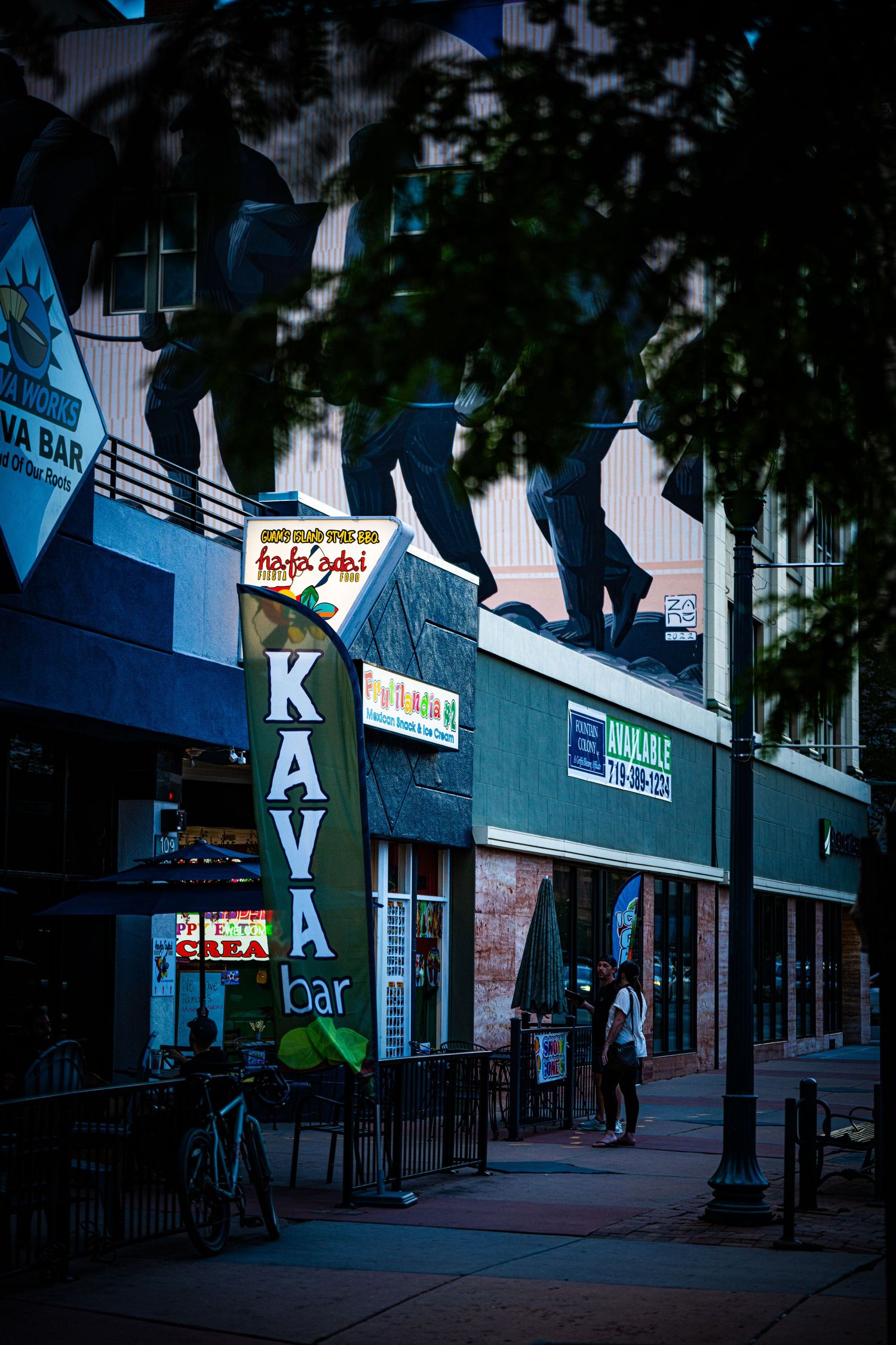 Kava Bar sign on a dimly lit urban street at dusk with storefronts and pedestrians in the background.