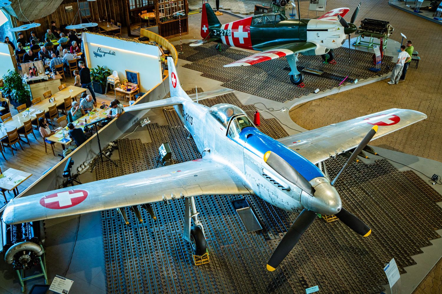 Vintage silver Swiss Air Force fighter plane with red cross markings on display in an aviation museum with visitors and other aircraft.
