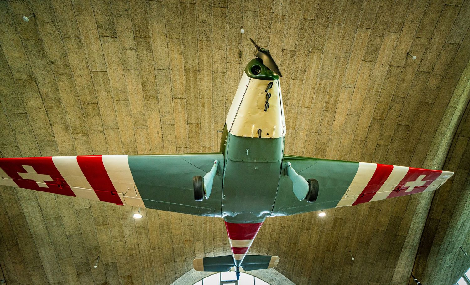 Vintage Swiss military aircraft with red and white wings and cross markings suspended from a wooden ceiling in a museum.