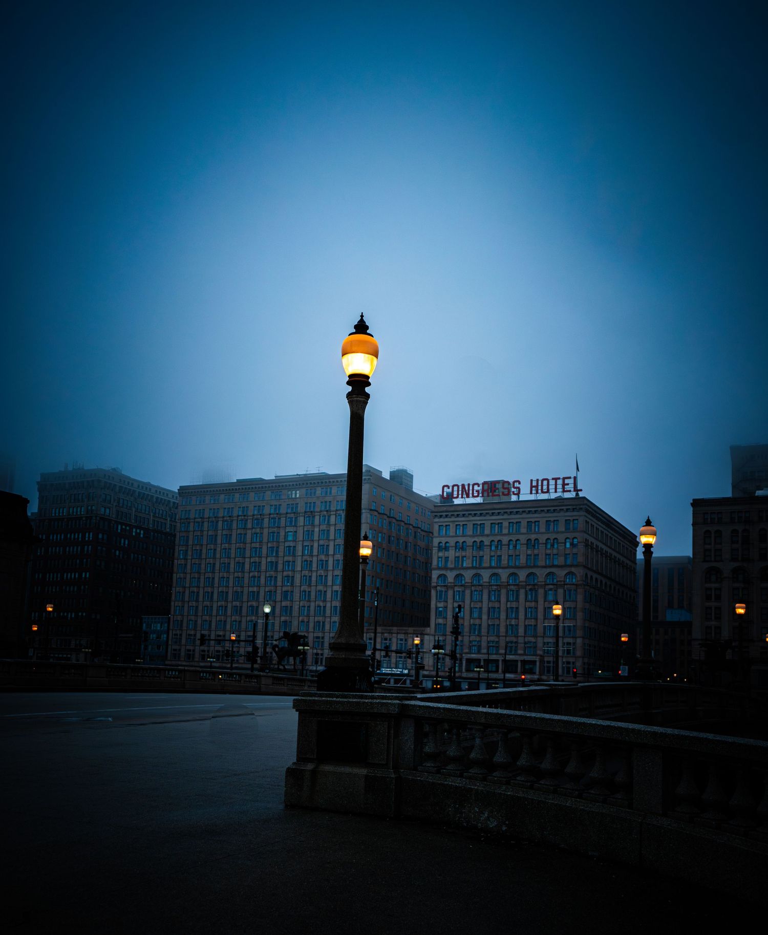 Moody cityscape with illuminated lampposts silhouetted against a dusky blue sky.