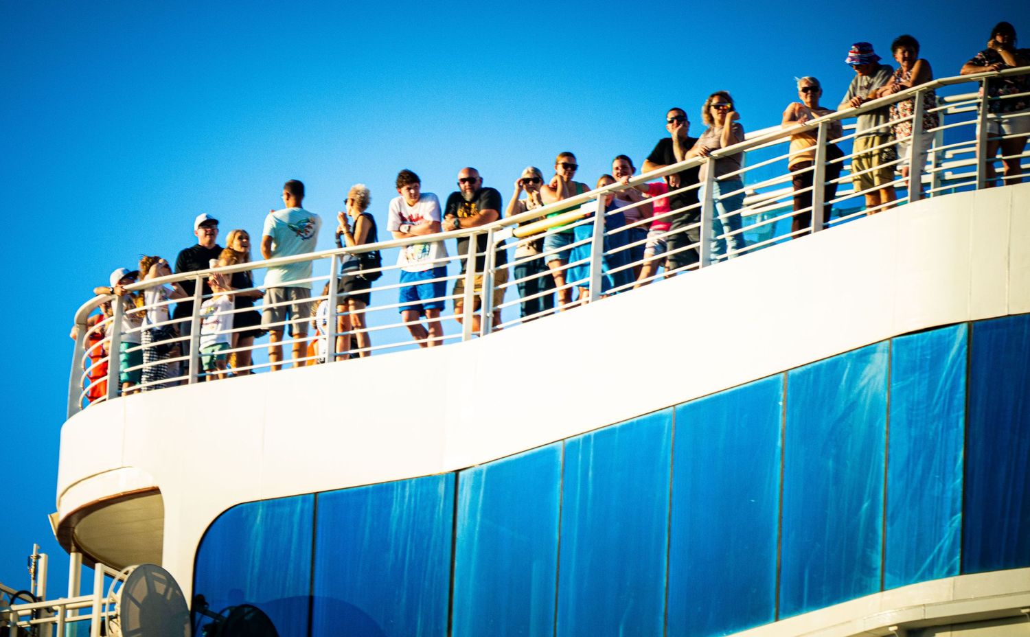 Passengers line the upper deck railings of a cruise ship against a bright blue sky on a sunny day.