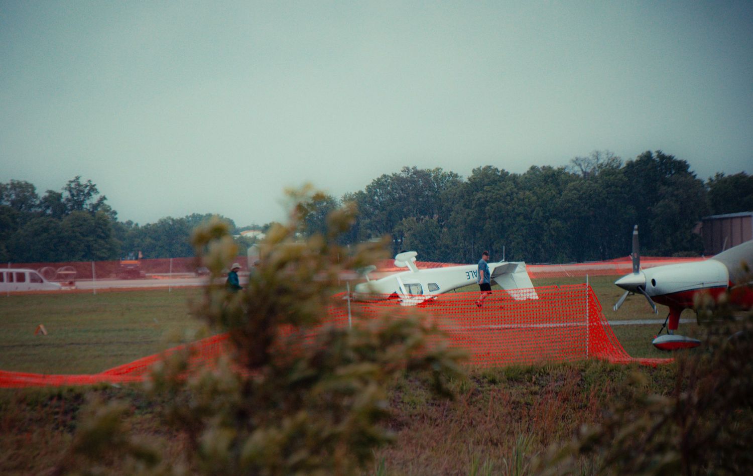 A small aircraft crashed and overturned on a red dirt runway with trees visible in the background.